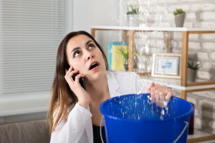 Worried Woman Calling Plumber While Collecting Water Droplets Leaking From Ceiling At Home