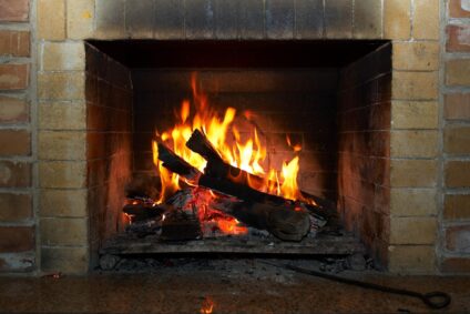 A burning fire in a home fireplace with visible soot and smoke residue on the surrounding brick hearth.